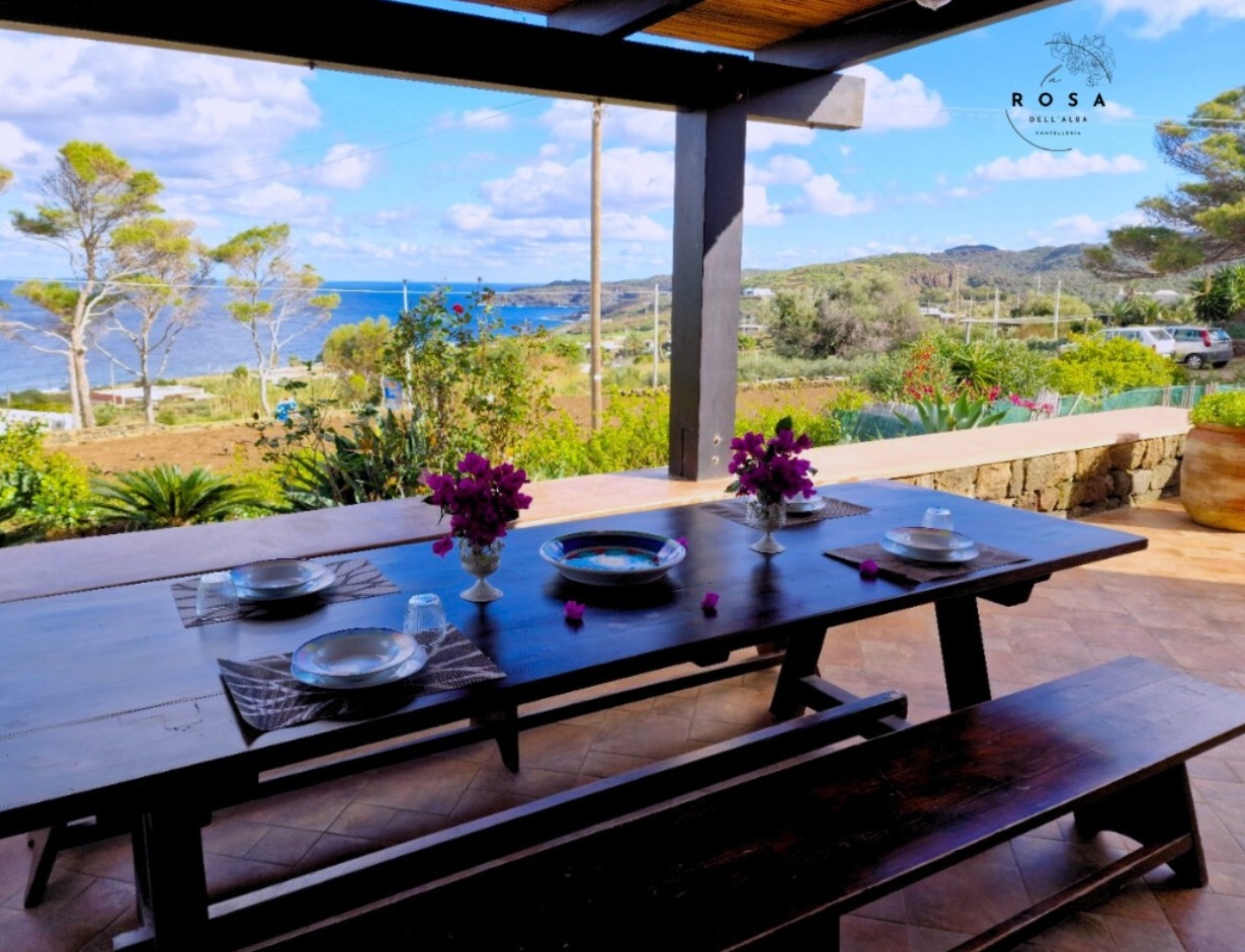 Tavolo da pranzo all’aperto sotto la veranda coperta, con vista sul mare e sulla costa di Pantelleria